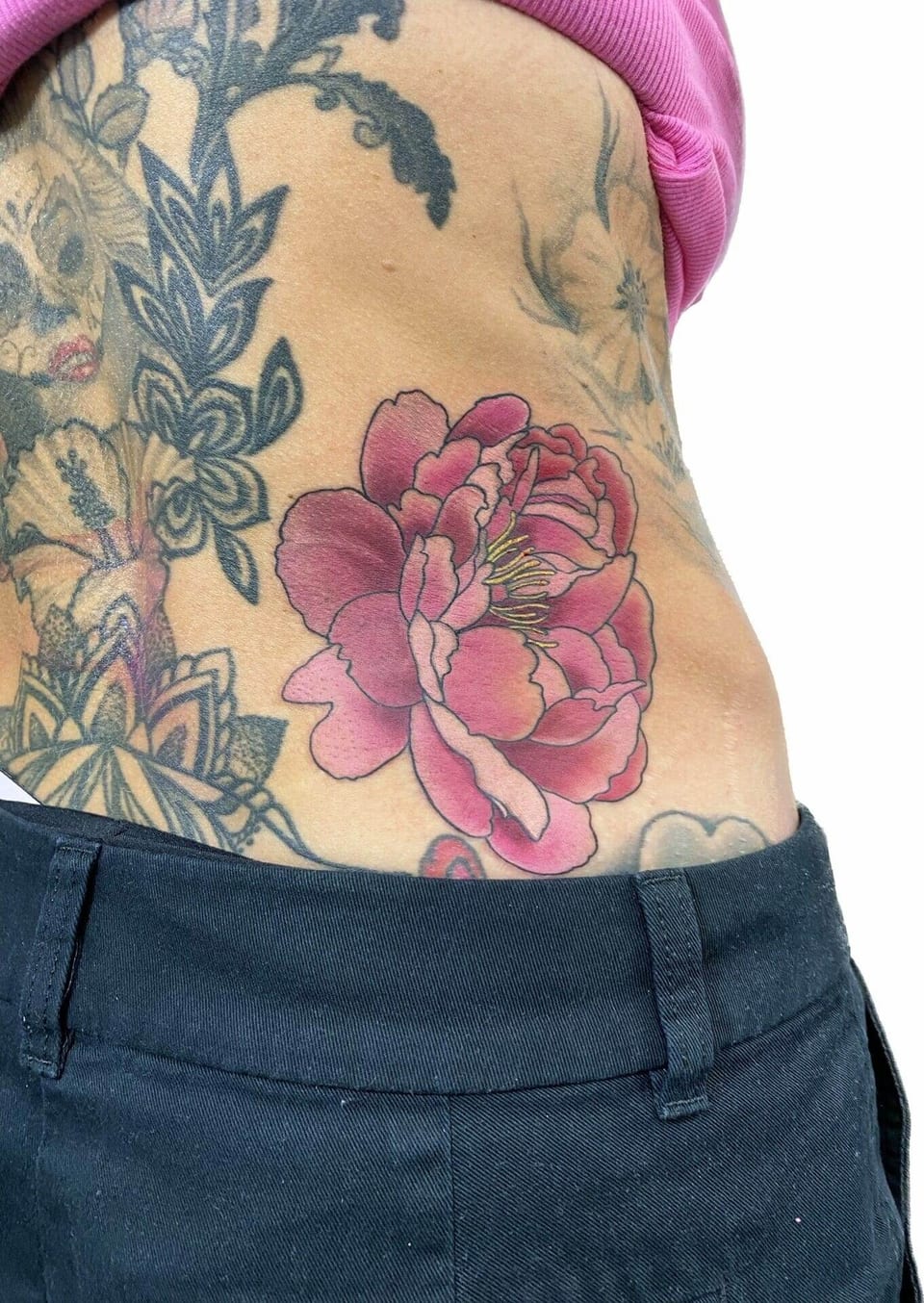 A neo-traditional pink peony with layered petals appears at the waist among black floral tattoos.