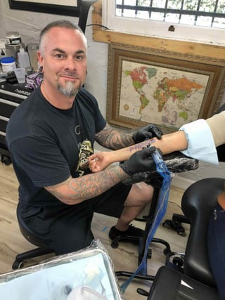 Seth Reynolds tattooing a client's arm beside a framed world map at his station