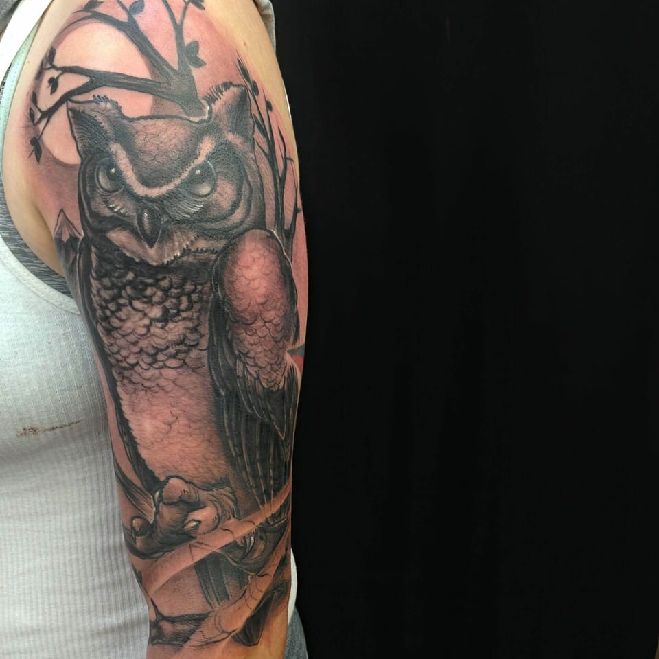 A black and grey realism owl perches on branches against a moon on the upper arm.