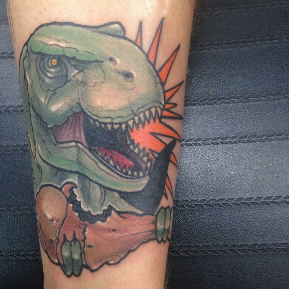 A neo-traditional green dinosaur bares its teeth above a bone against orange rays on a cropped limb.