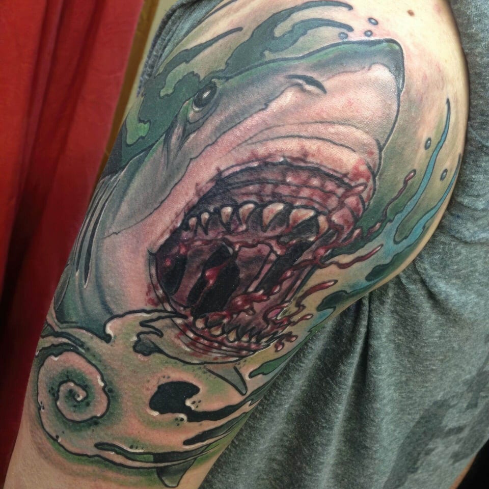 A shark opens its jaws above a spiral shell amid green and blue swirls on an upper arm.
