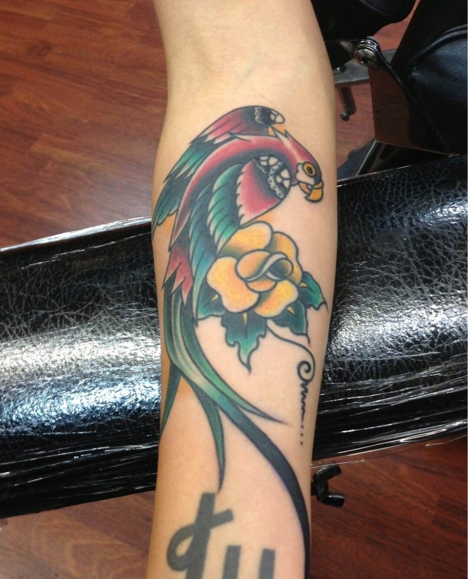 A traditional red and green parrot curves around a yellow rose on the forearm.