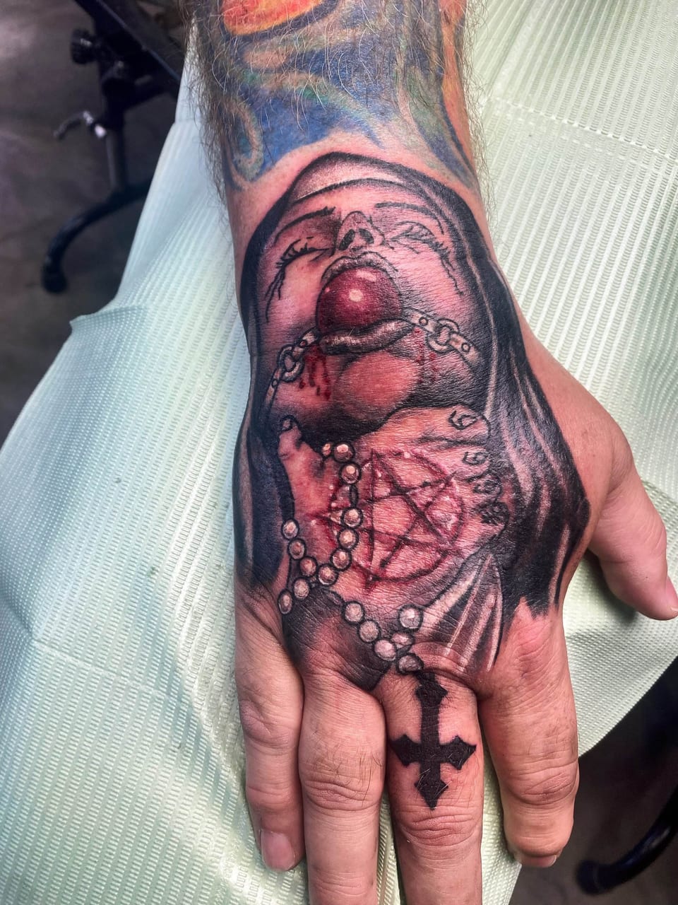 A black and grey illustrative veiled nun with a red ball gag covers the hand above an inverted cross on a finger.