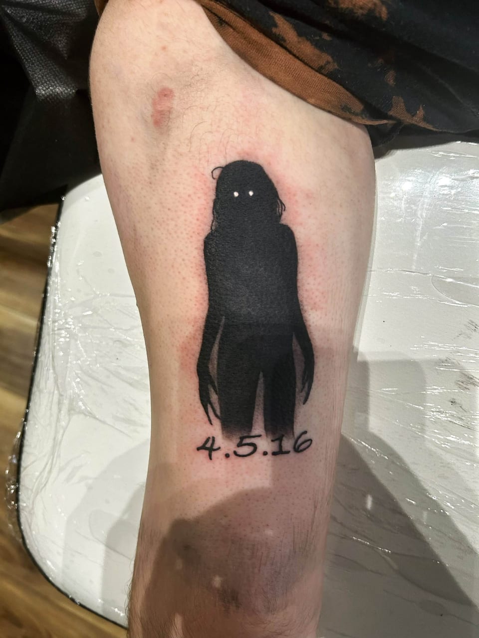 A blackwork figure with white eyes stands above a handwritten date on the upper arm.