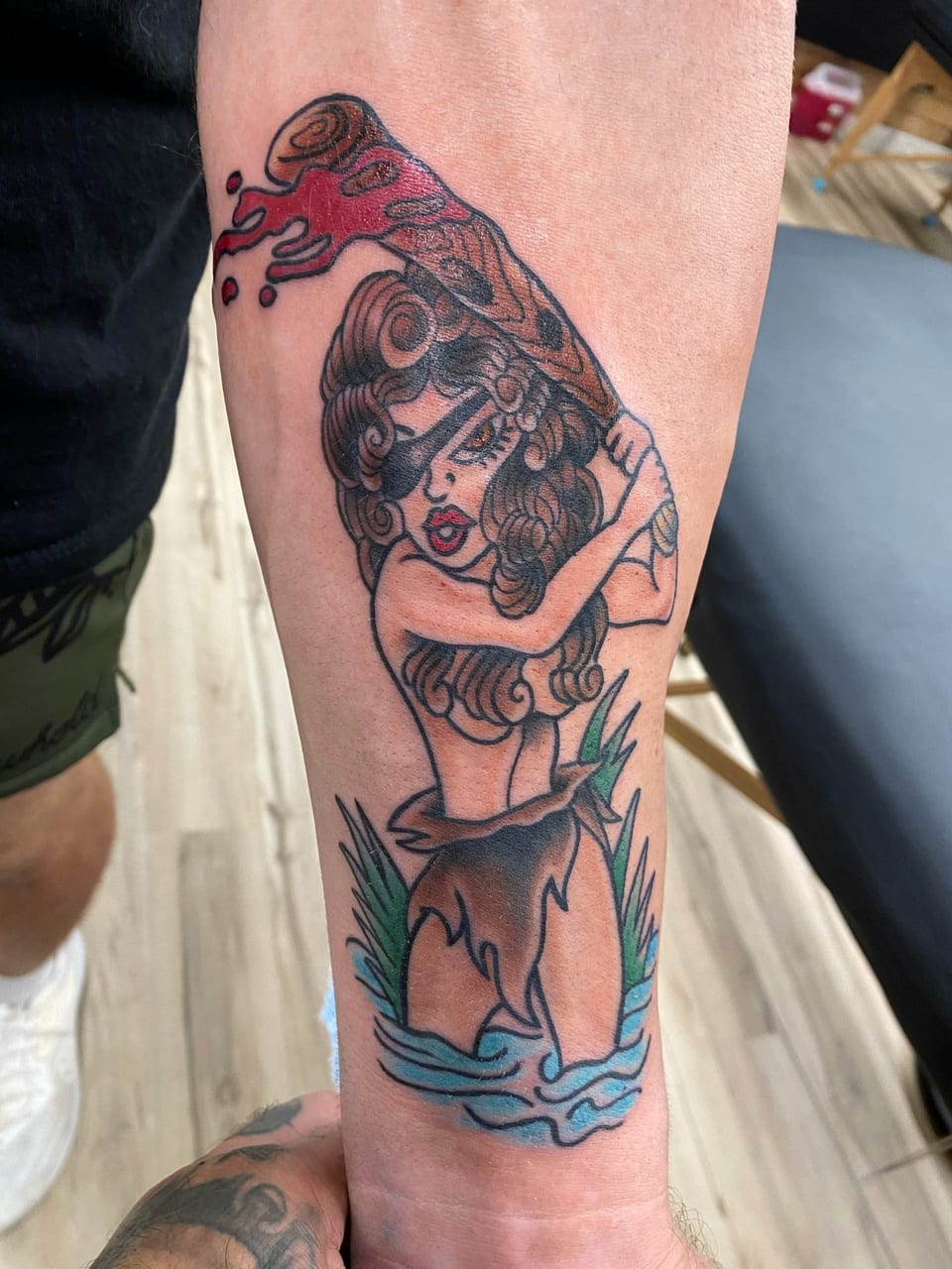 A traditional woman in a leaf skirt raises a bloody club while wading in water on a forearm.