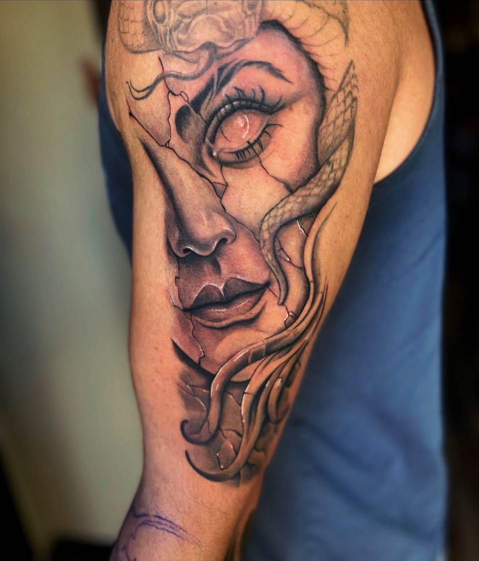 A realistic black and grey woman's face with stone-like cracks and a snake covers an upper arm.