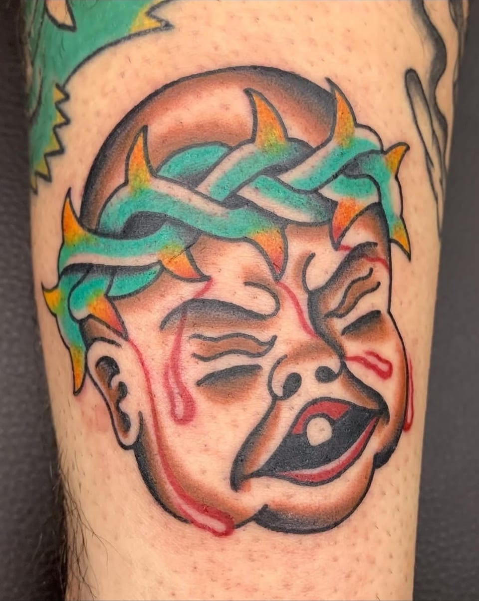 A crying face tattoo wears a green thorn crown with yellow tips.