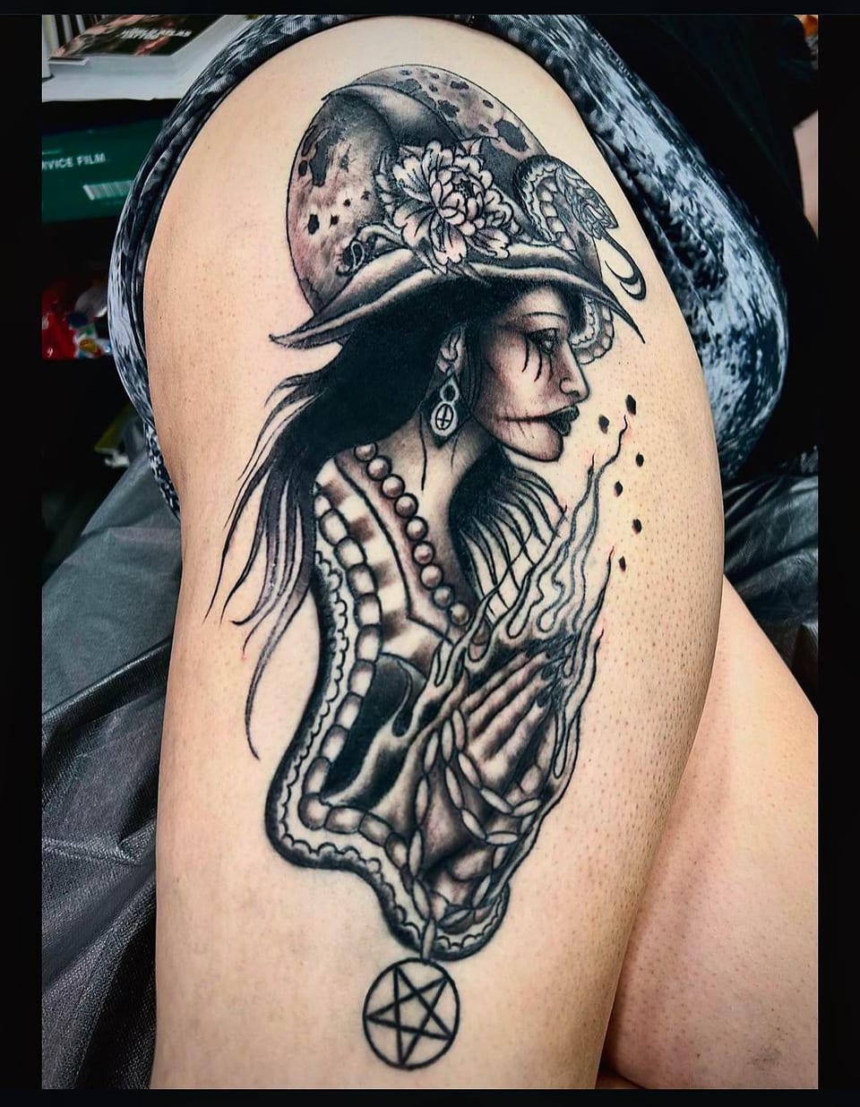 A black and grey illustrative witch with a pointed hat, snake and flames covers the thigh.