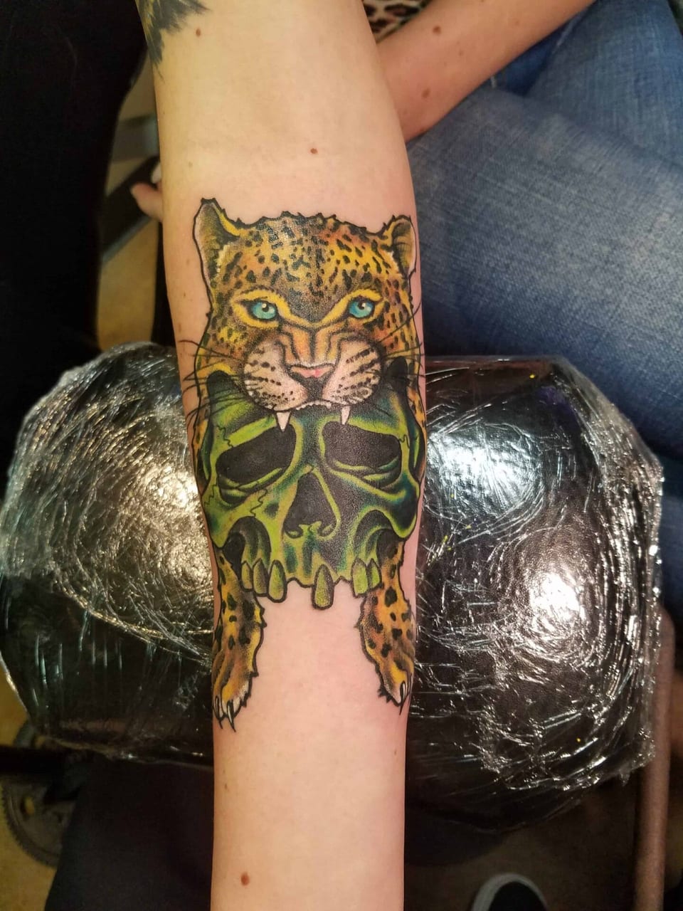A color illustrative leopard grips a green skull on a forearm.
