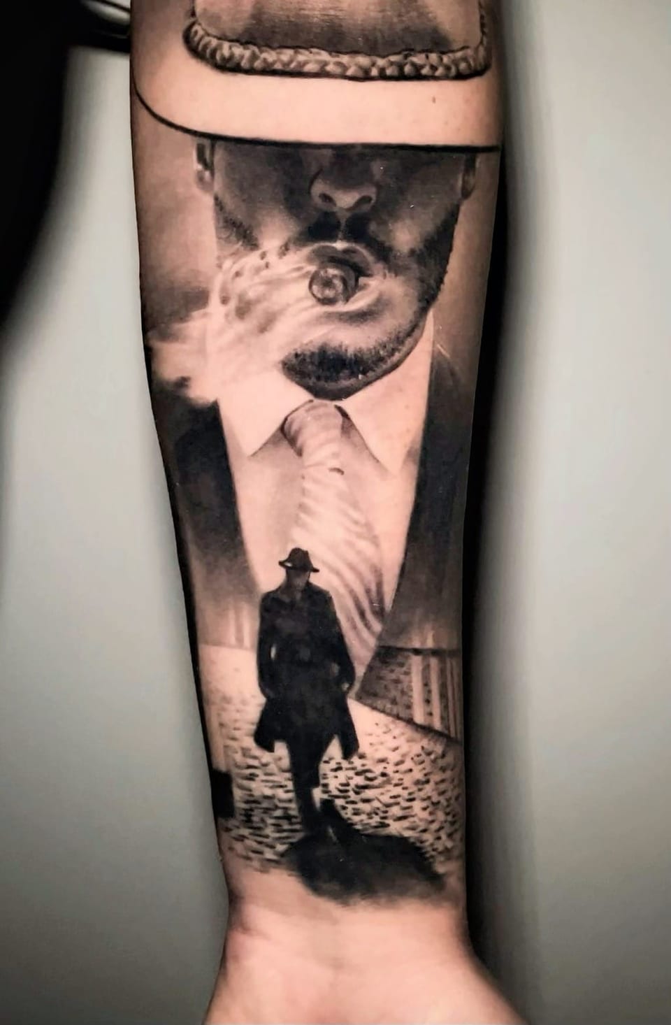 A black and grey realism man with a hat and cigar looms above a walking figure on a forearm.