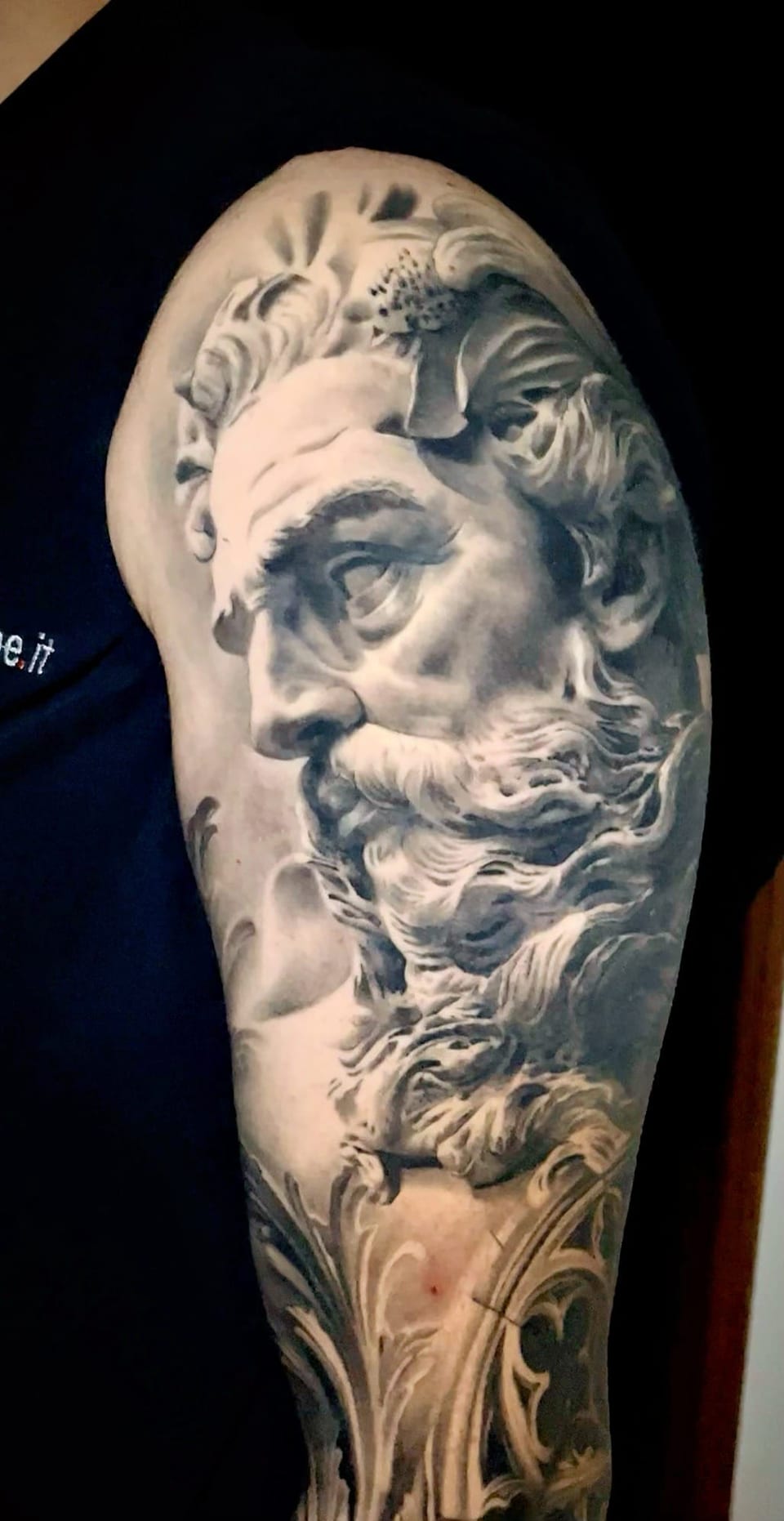 A black and grey realism bearded statue with flowing ornamental shapes covers an upper arm.