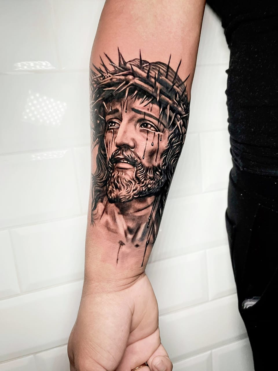 A black and grey realism portrait of Jesus with tears and a crown of thorns covers a forearm.