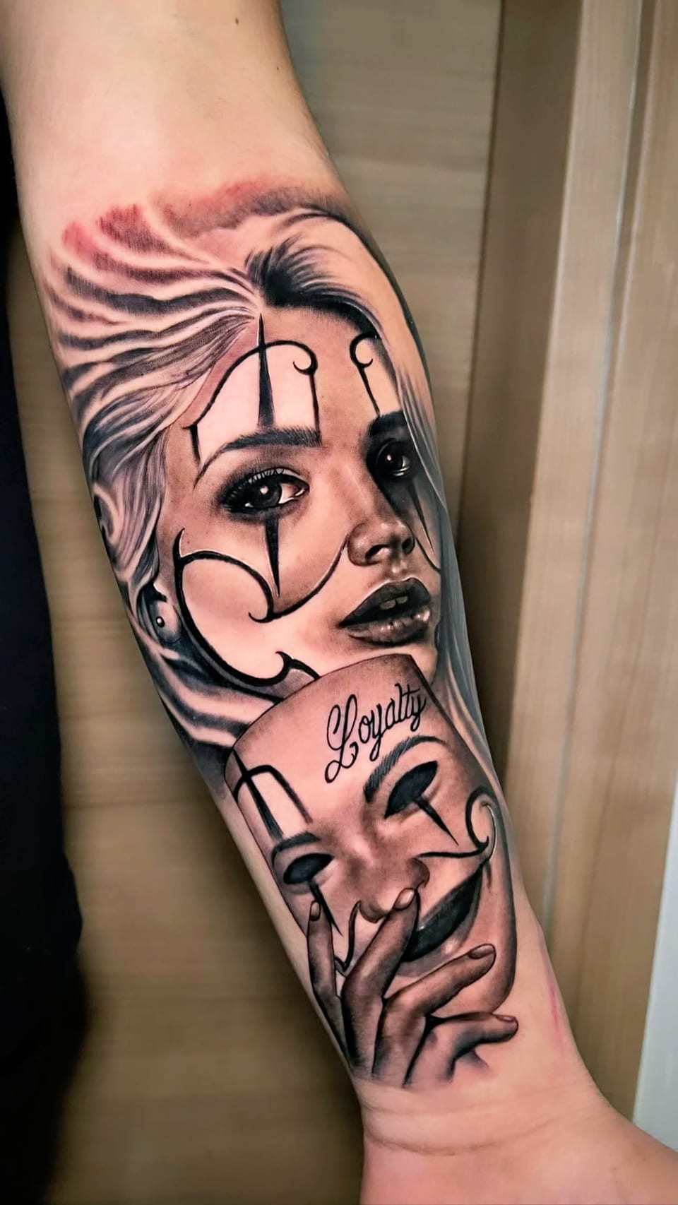 A black and grey realism woman with painted tear lines holds a smiling mask marked Loyalty on a forearm, against wood.