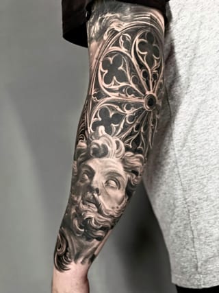 A black and grey realism bearded statue sits beneath an ornate circular window on a forearm.