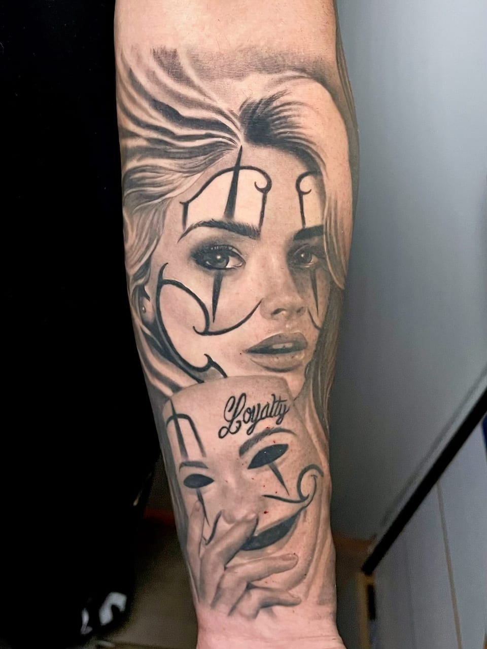 A black and grey realism woman's face with clown-style lines sits above a "Loyalty" mask on the forearm.
