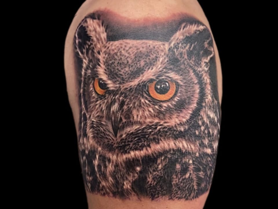 A black and grey realism owl with orange eyes covers an upper arm.