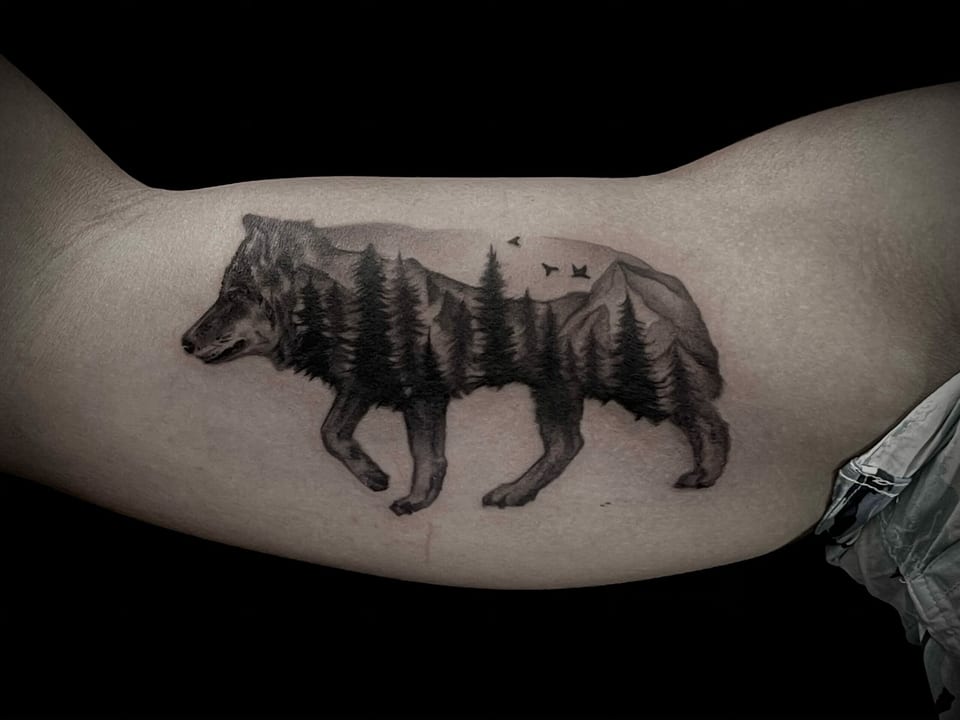 A black and grey illustrative wolf silhouette filled with forest and mountains covers an inner upper arm.