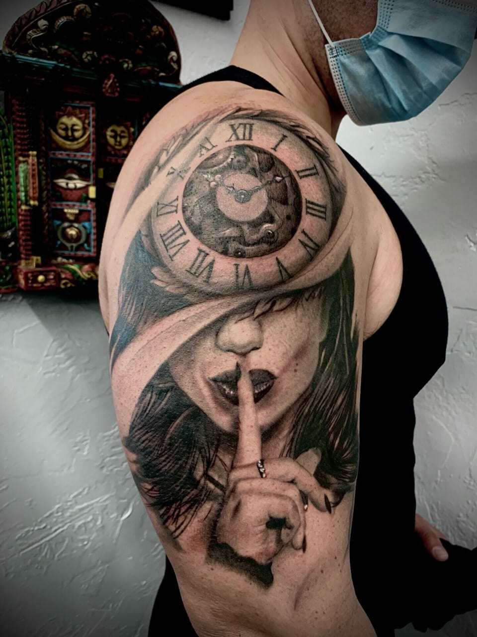 A black and grey realism woman making a shushing gesture beneath a clock covers an upper arm.