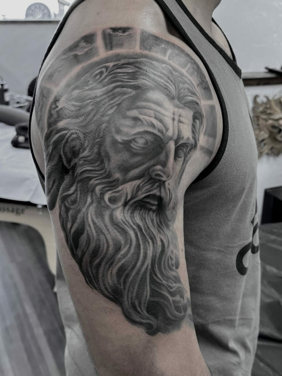A black and grey realism bearded classical statue beneath a segmented halo covers an upper arm.