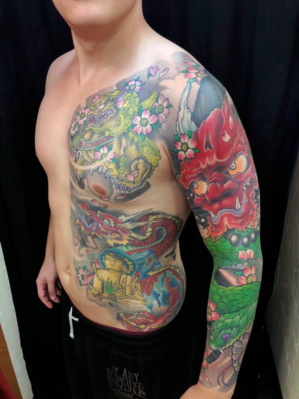 Color Japanese dragon, guardian lion, mask and blossoms span an arm and the side of a torso.