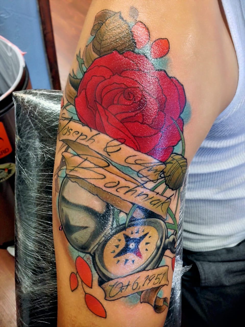 A color illustrative red rose, pocket watch and scroll banners cover an upper arm.