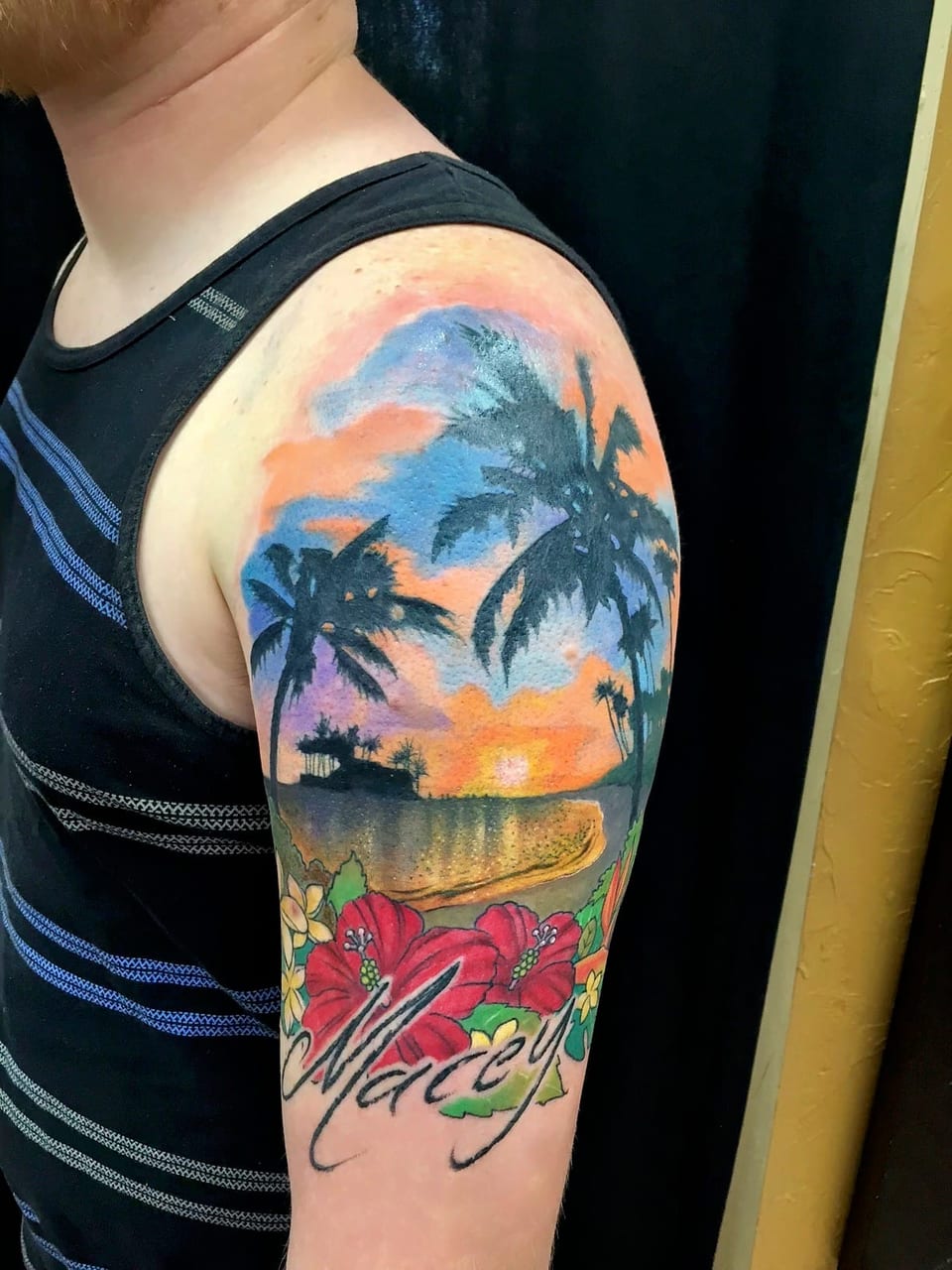 Color realism tattoo of a palm-lined sunset beach and hibiscus flowers covers the upper arm.