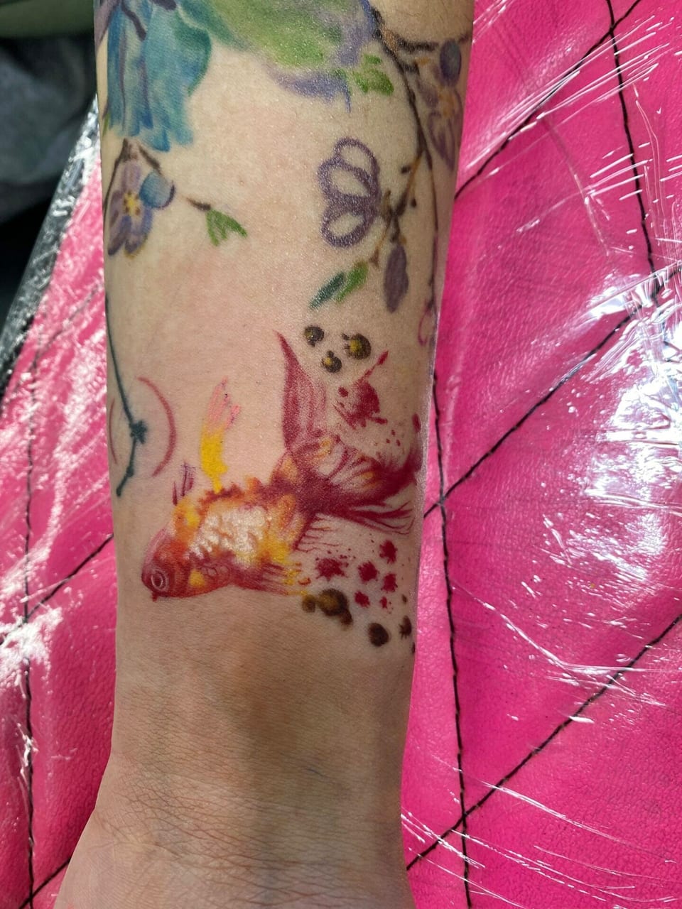 A red and yellow watercolor fish swims amid scattered spots on the forearm.