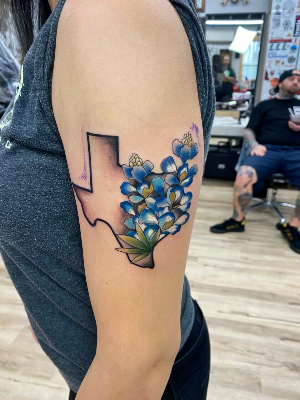 Illustrative color tattoo combines a Texas outline and blue flowers on the upper arm.