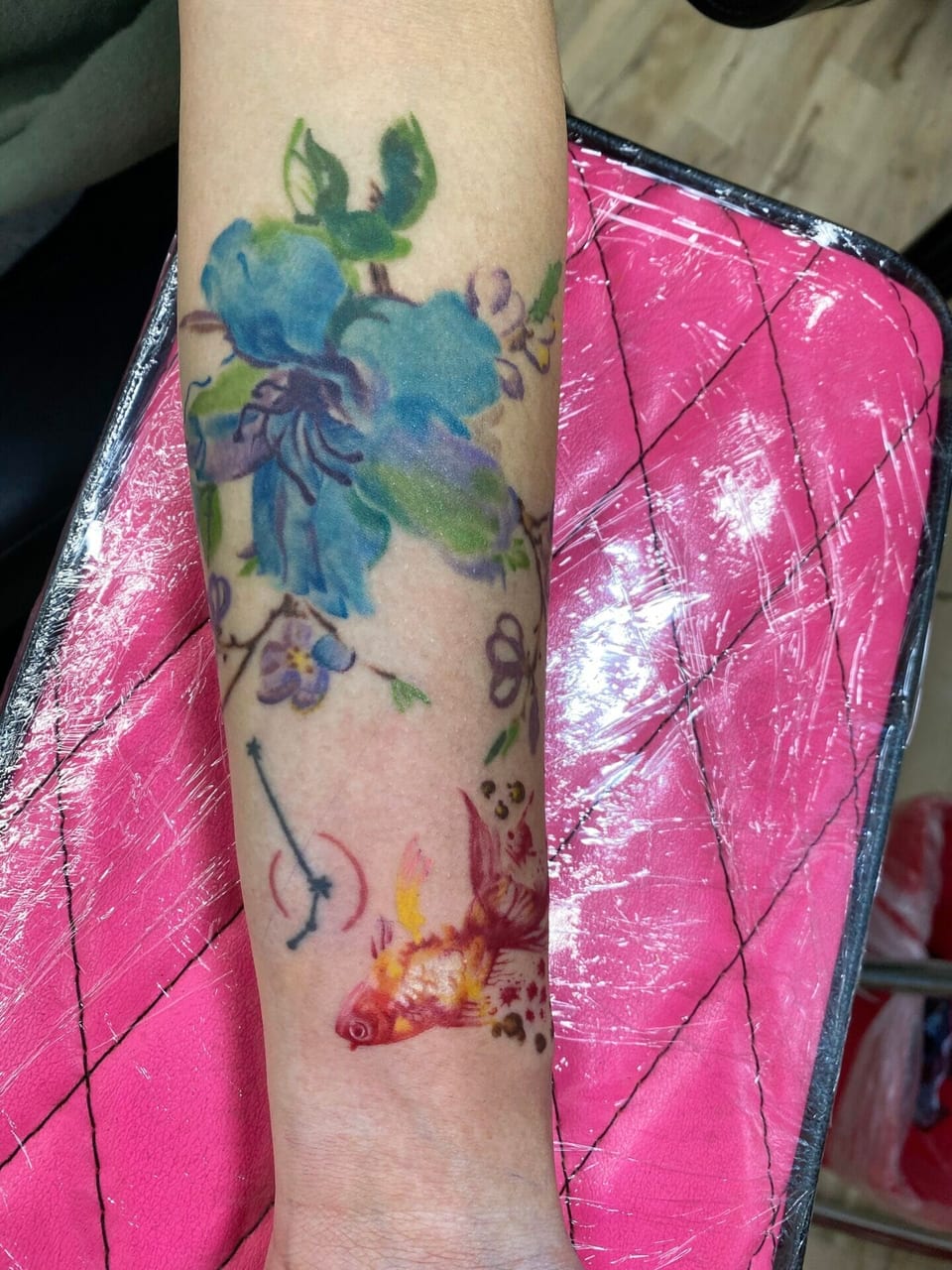 Blue and green watercolor flowers appear above a red and yellow fish on a forearm.