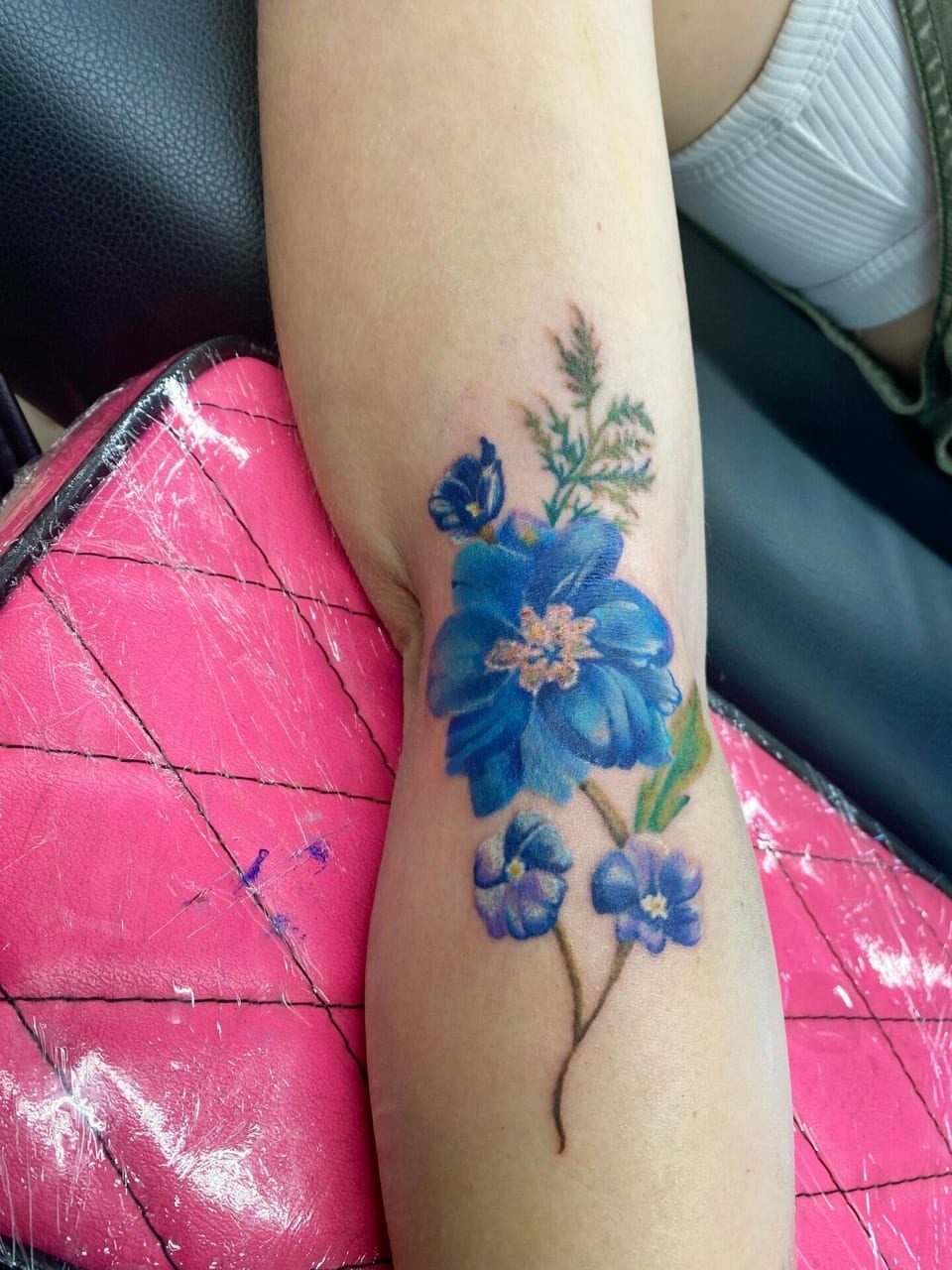 Color realism tattoo of blue flowers with green foliage sits on the inner forearm near the elbow.