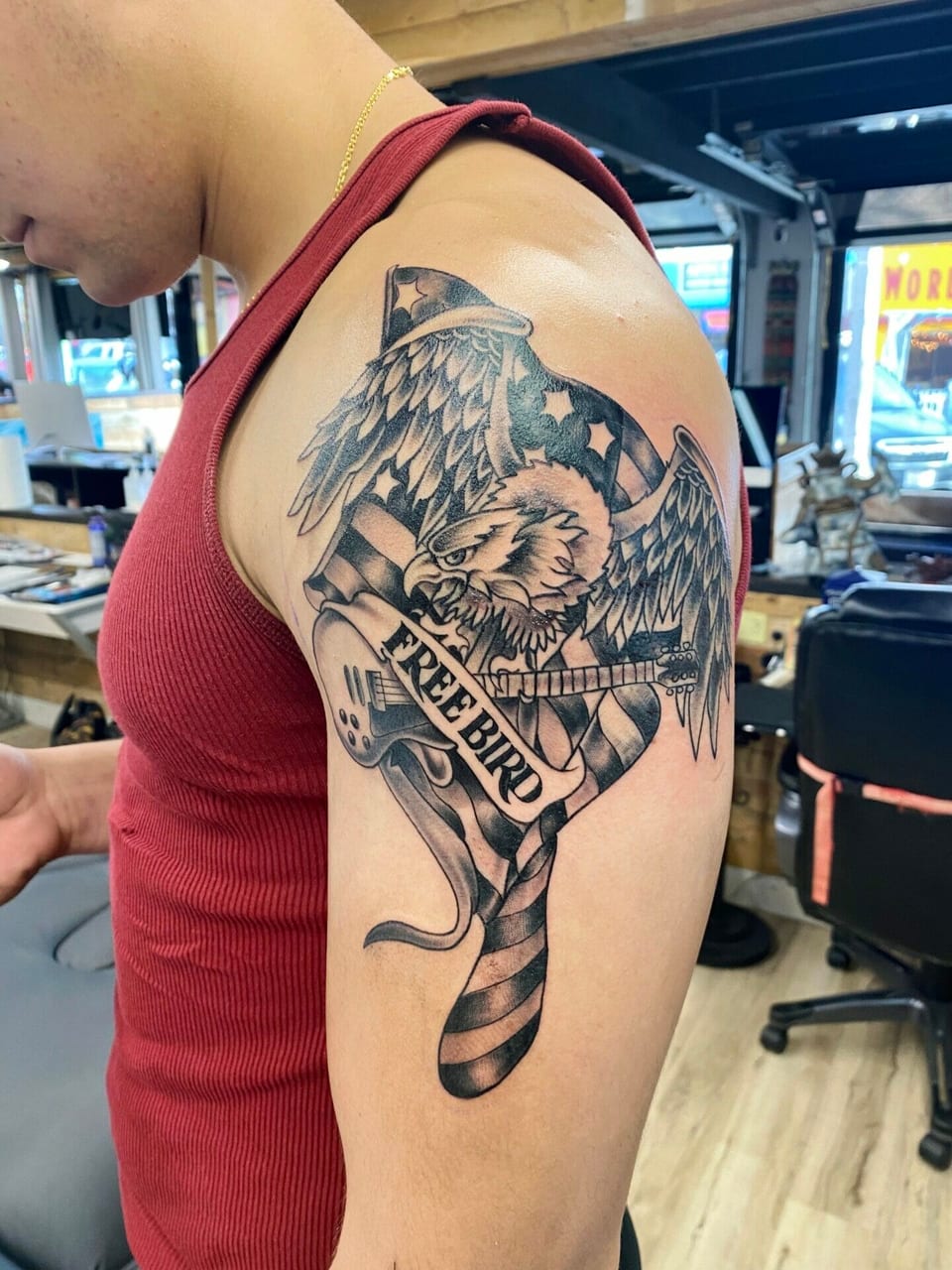 Black and grey illustrative tattoo of an eagle, guitar, flag and 'Free bird' banner covers the upper arm.