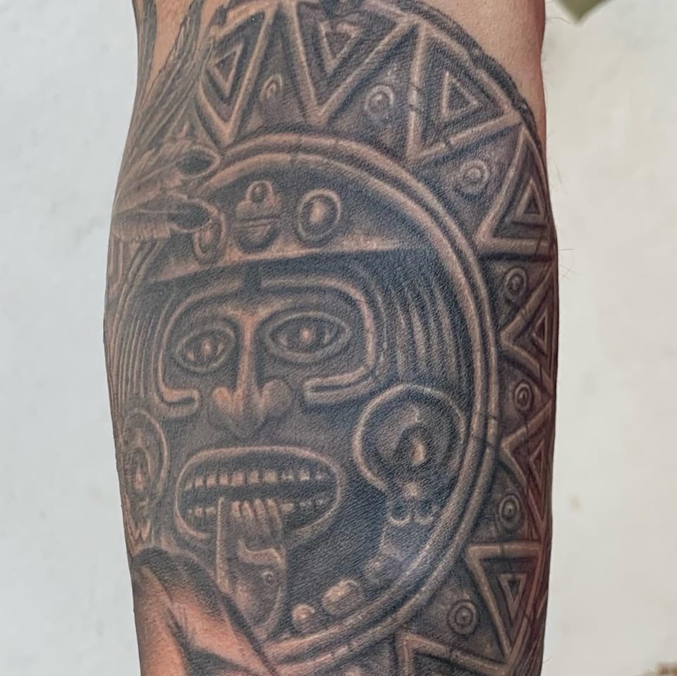 A realistic black and grey carved sun-stone face with a geometric border covers a tightly cropped arm.