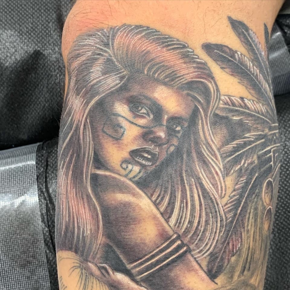 A realistic black and grey woman with face paint appears beside feathers in a tightly cropped arm tattoo.