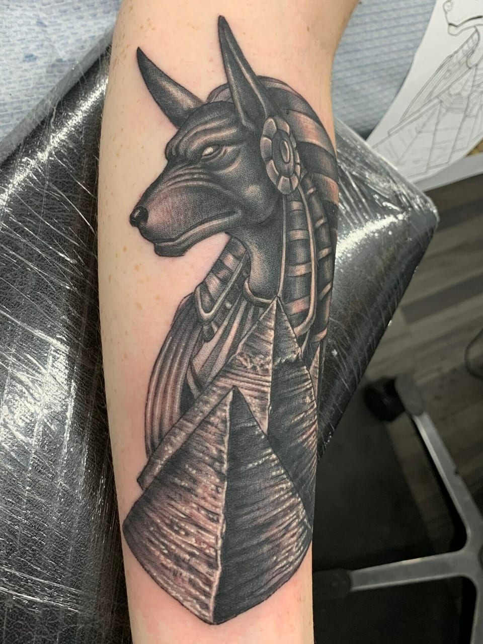 Black and grey realism Anubis tattoo with a striped headdress and pyramids covers the forearm.