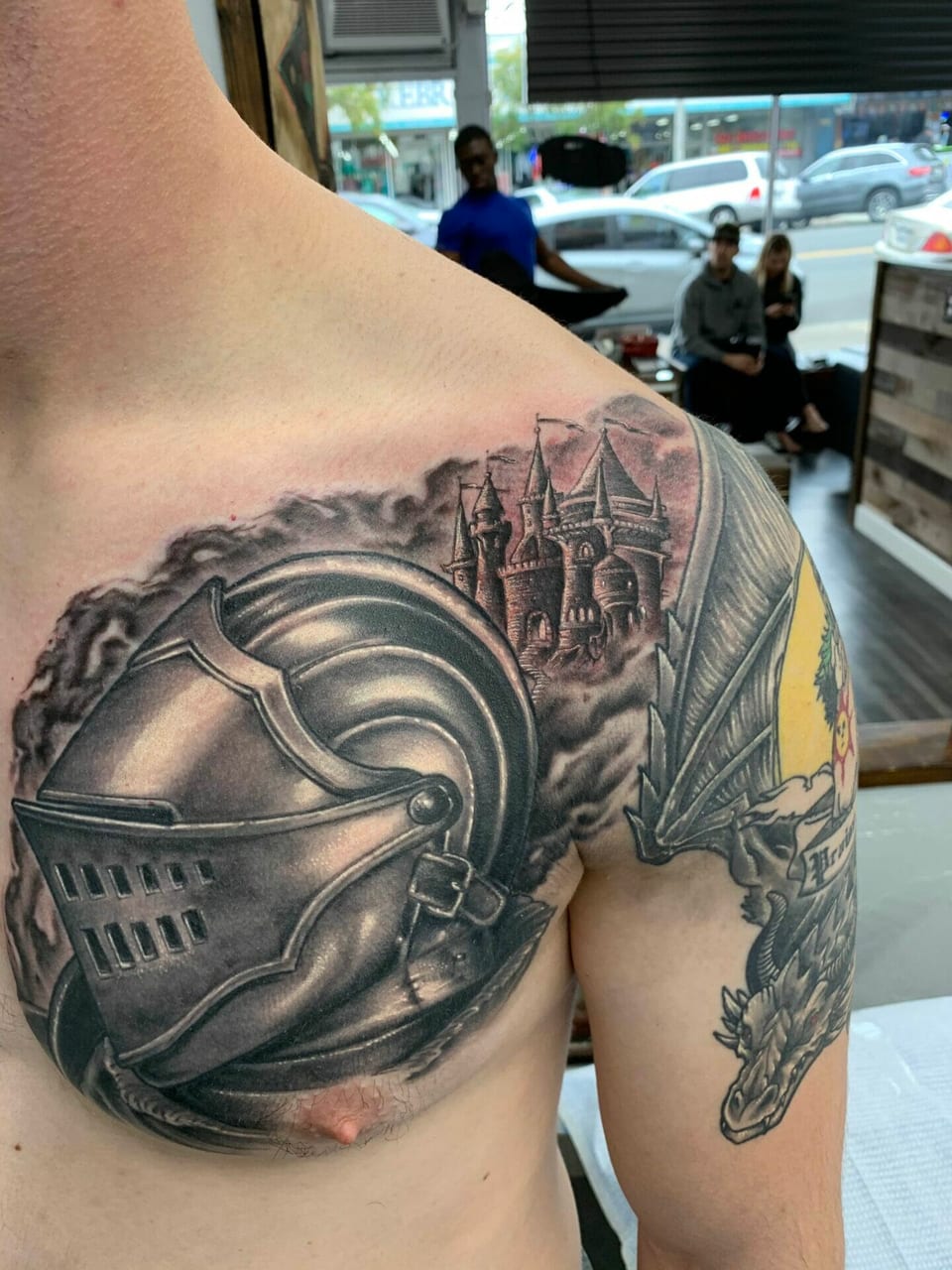 Black and grey realism knight's helmet and castle tattoos cover the chest and shoulder beside a dragon.