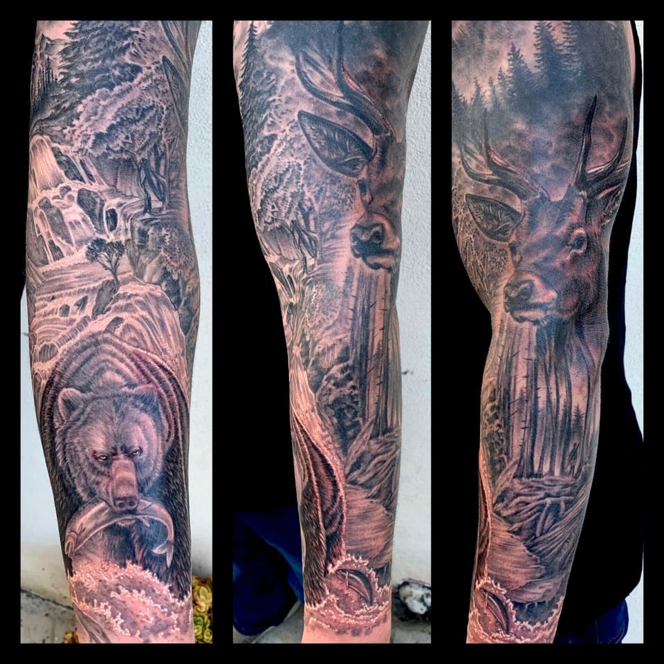 Black and grey realism forest sleeve with a bear, stag and waterfall covers the arm in several views.
