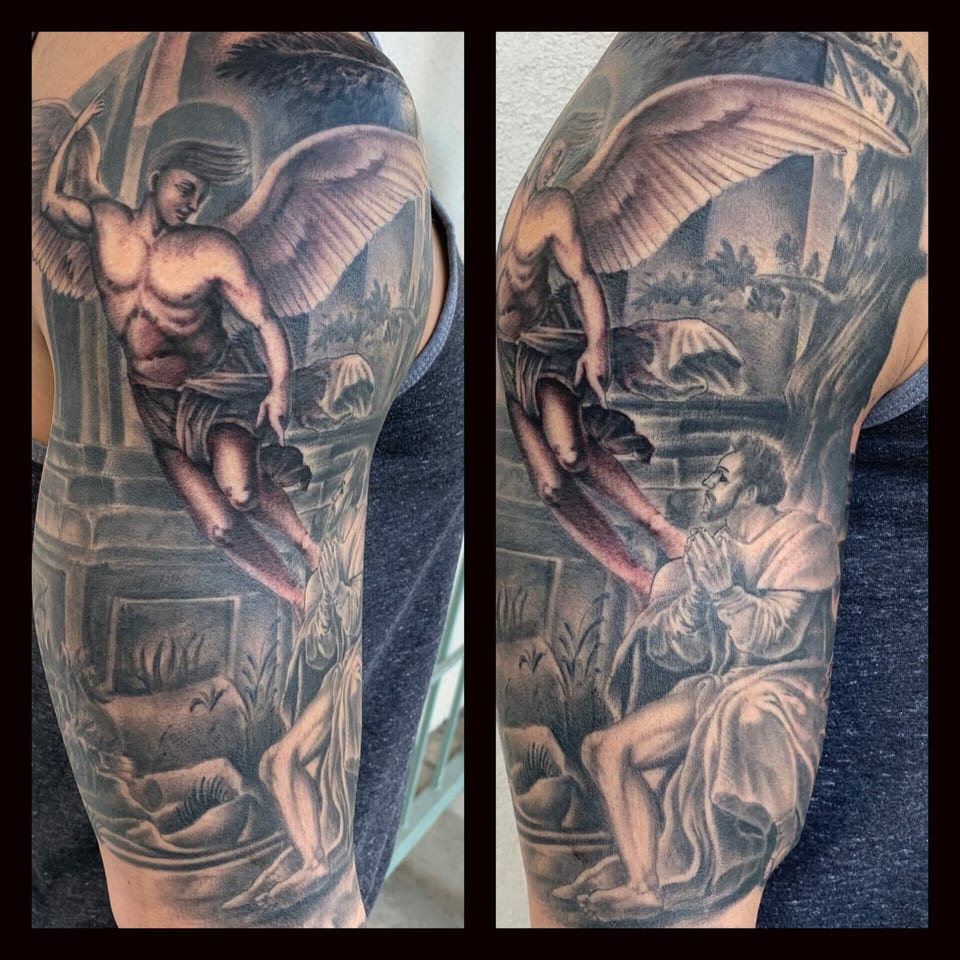 Black and grey realism angel and kneeling man cover the upper arm in a garden scene.