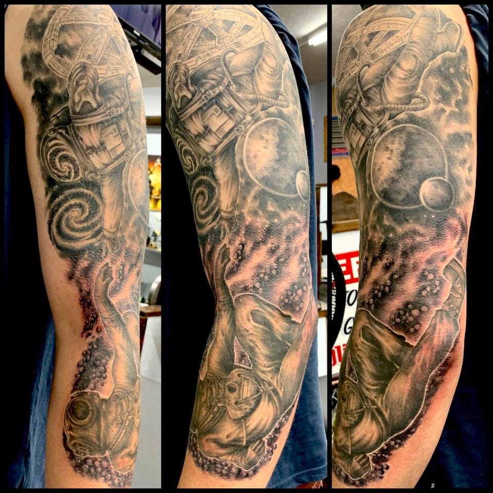 A black and grey realism arm sleeve combines a space station, astronaut, planets and diver in a collage.