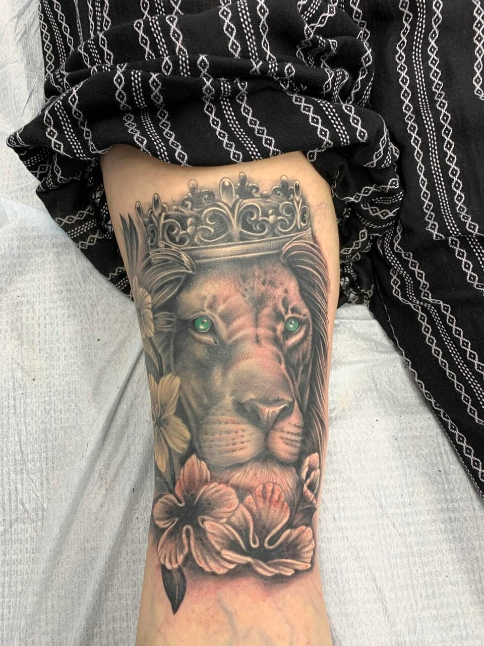 Black and grey realism crowned lion with green eyes and flowers covers the forearm.