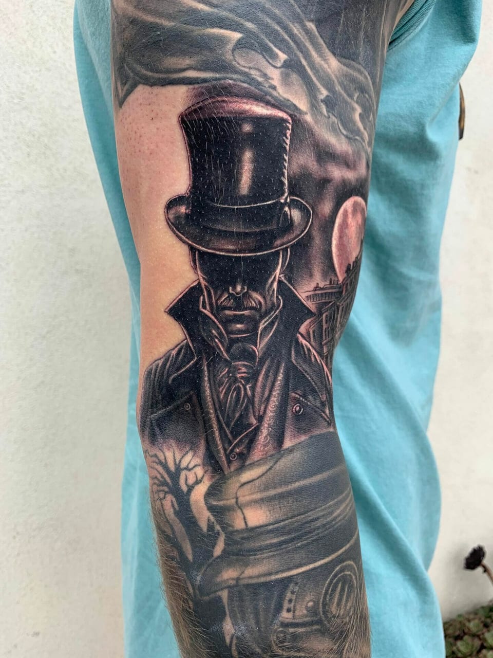 Black and grey realism tattoo of a man in a top hat and long coat covers the arm.