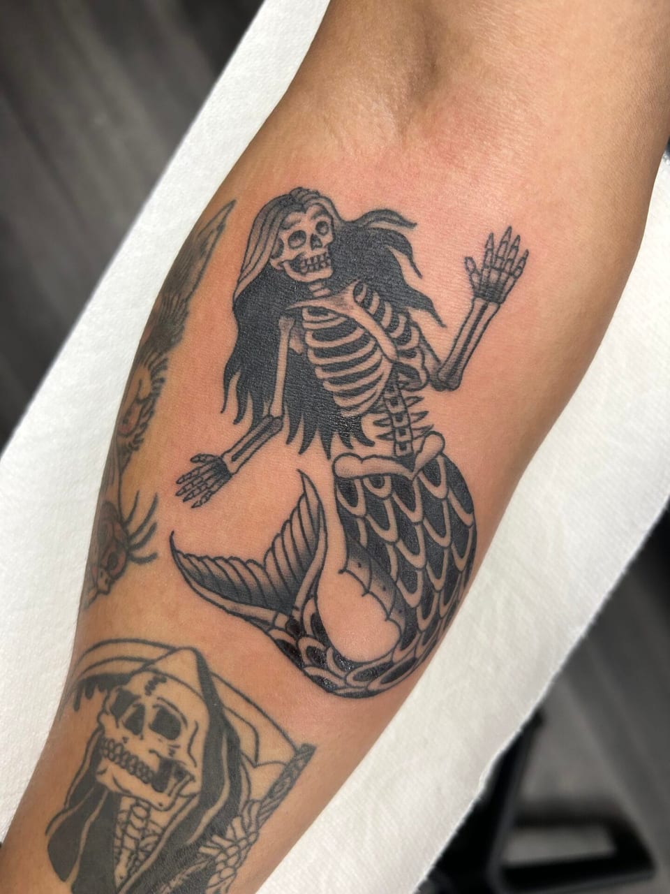 Black and grey traditional skeletal mermaid with long hair sits on the forearm.