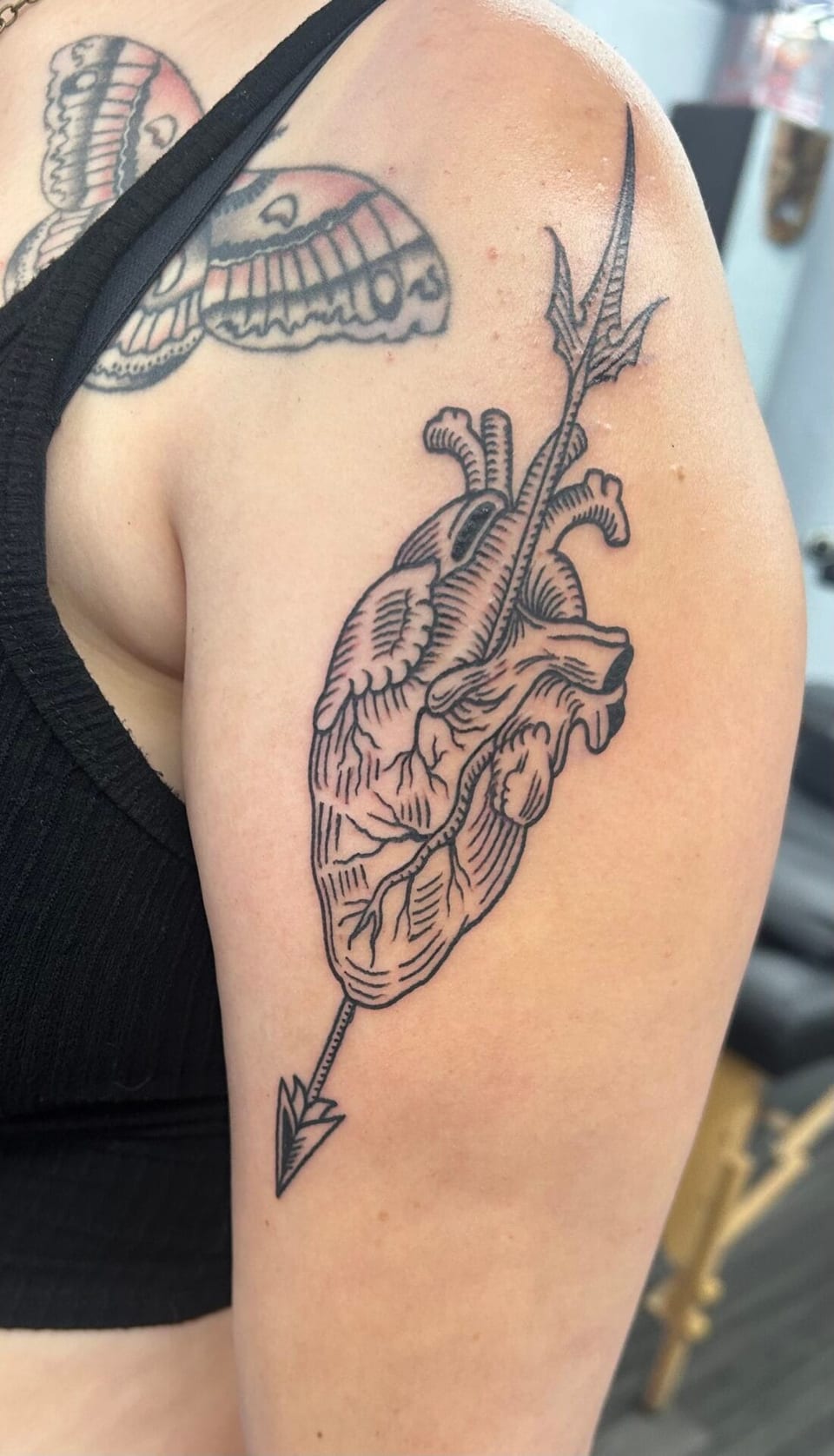 Black illustrative anatomical heart pierced by an arrow sits on the upper arm.