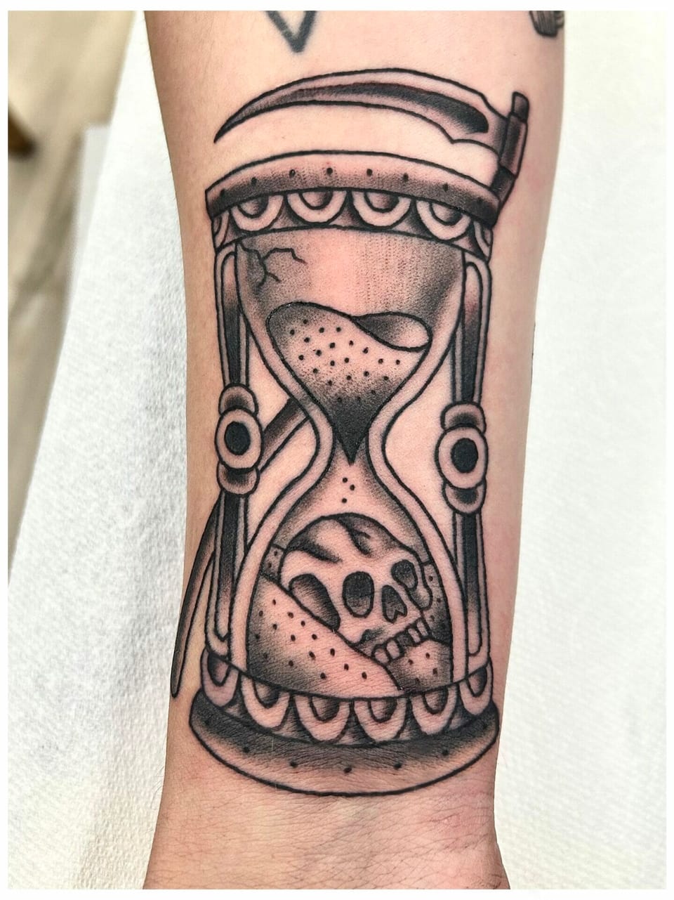 Black and grey traditional hourglass with a skull and scythe sits on the forearm.