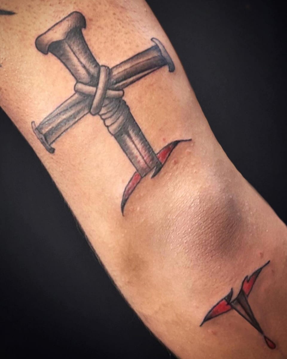 Black and grey illustrative crossed nails bound with cord sit above the elbow with red torn-skin accents.