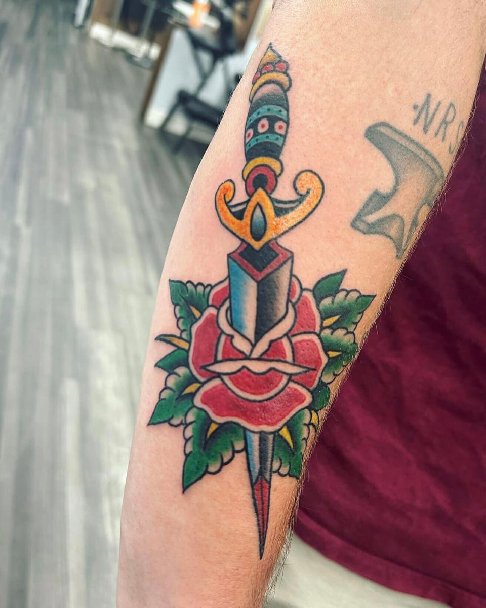 Traditional color dagger piercing a red rose sits on the forearm.