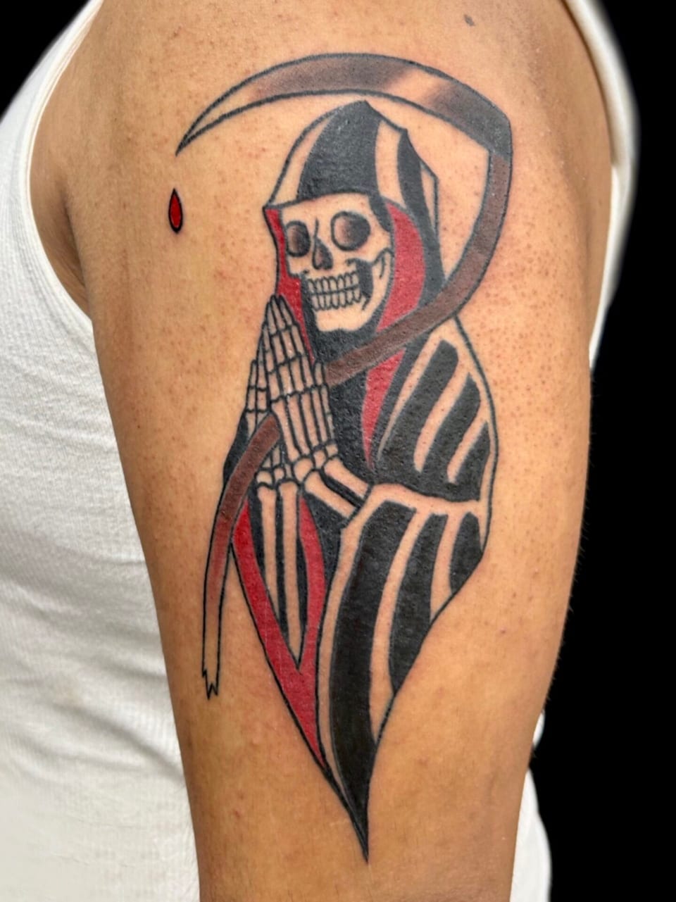 Traditional black and grey grim reaper with praying hands, a scythe and red accents sits on the upper arm.