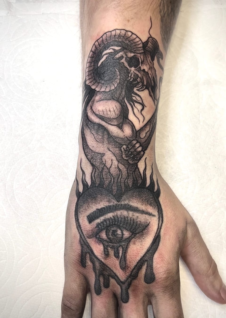 Black and grey illustrative horned figure above a weeping eye covers the wrist and back of the hand.