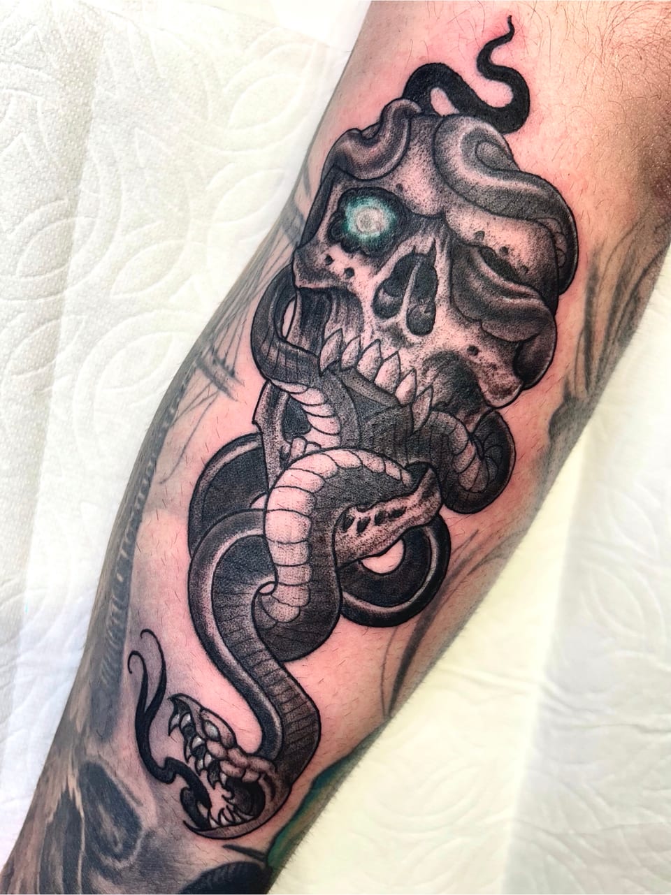 Black and grey illustrative skull entwined with a snake and a blue eye sits on the forearm.