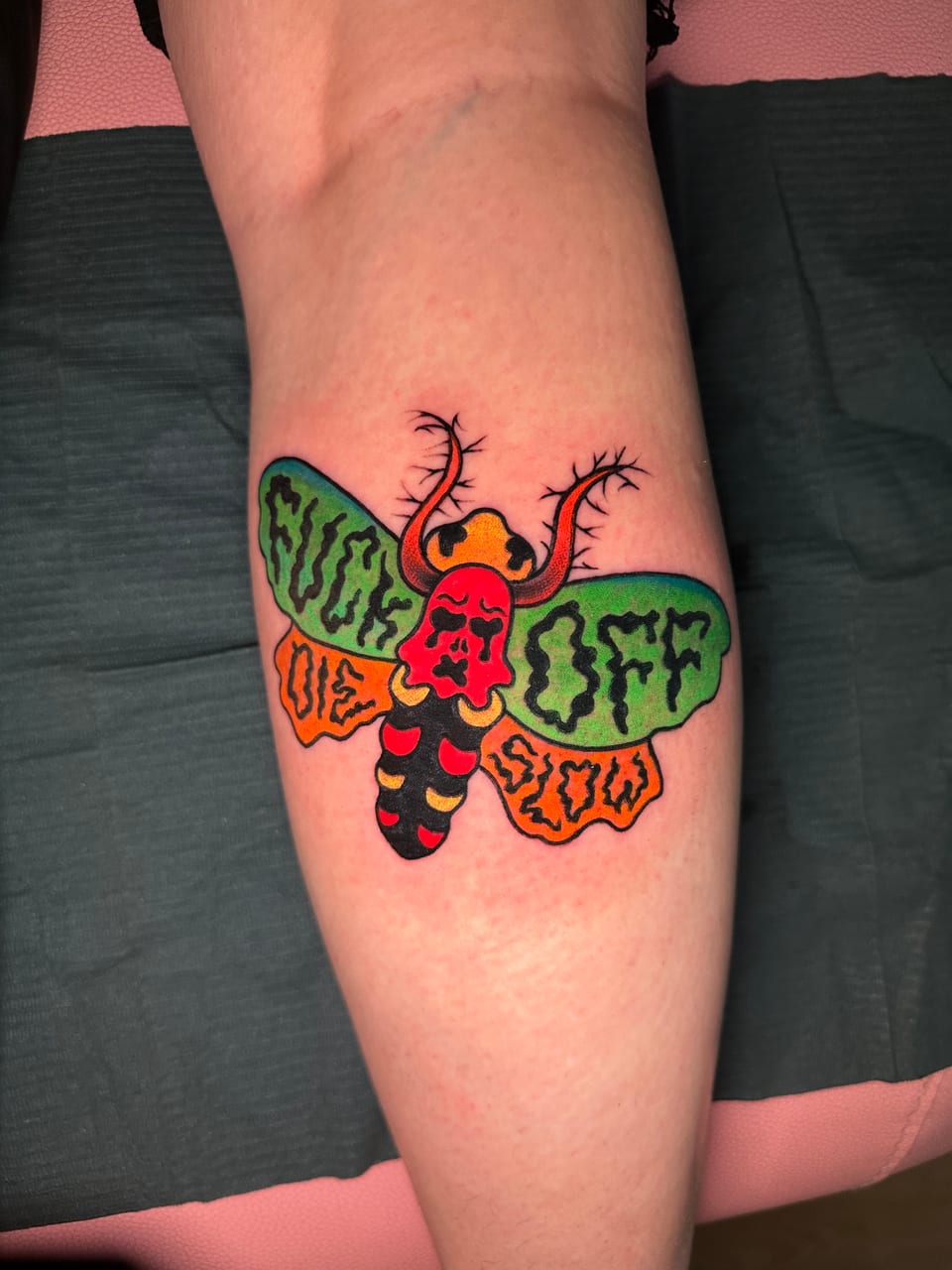 A red skull moth tattoo spreads green and orange wings with bold lettering across a calf.