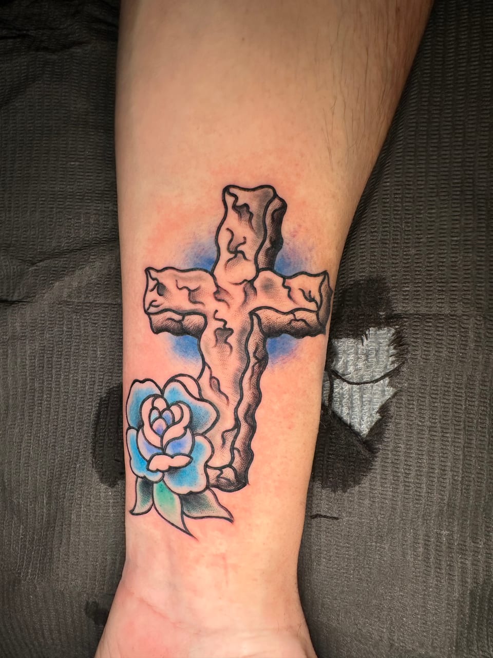 A shaded stone cross and blue rose tattoo fills the inner forearm.